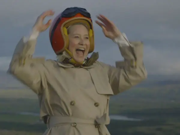 Glad Trine Dyrholm i pilotmössa och trenchcoat vinkar glatt. En scen från filmer med Trine Dyrholm.